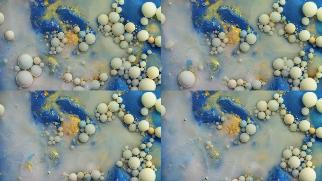 彩色金色丙烯酸涂料气泡散布在蓝色表面上，并混合在抽象设计中。闪闪发光的灰尘，油墨球滴和混合。奇妙的彩