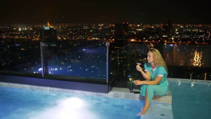这位漂亮的女商人花时间在豪华酒店里。女人在屋顶照明游泳池的边缘放松，喝鸡尾酒。在现代夜大城市休息旅行