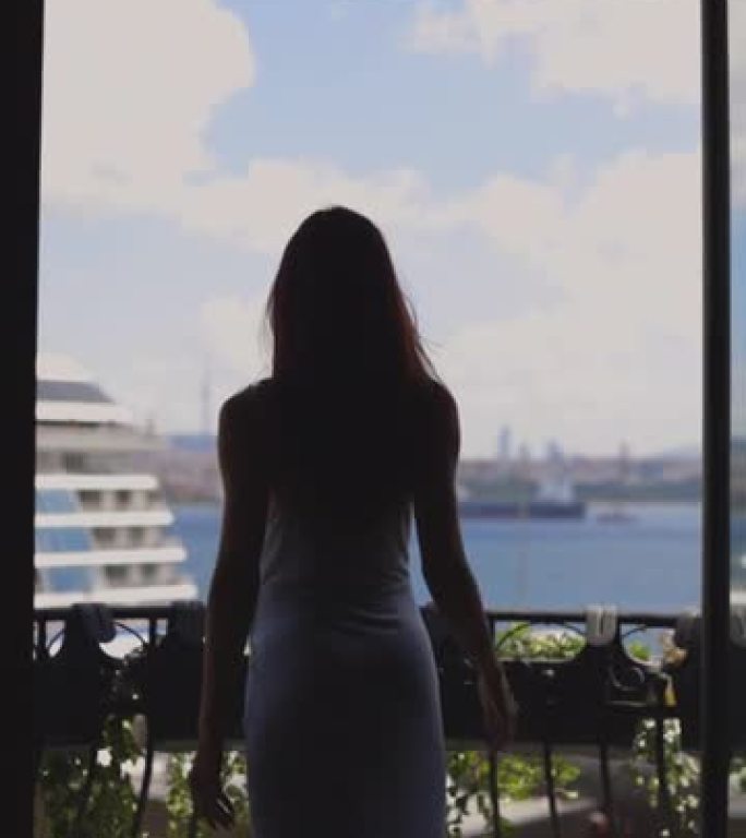 女人的剪影从酒店阳台看风景，一个穿着裙子的女人在酒店阳台上休息，看着大海和船的景色，女人打开窗帘