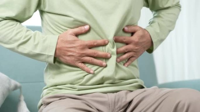 亚洲老人感到胃痛。年长的成熟男性在家坐在客厅的沙发上摸肚子。肠道问题医疗保健和医疗保险概念。