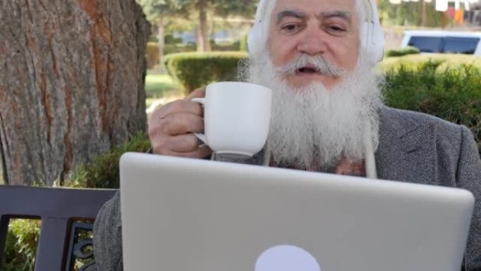 时尚聪明的高级男子拿着杯子喝热饮料，正在使用笔记本电脑和白色无线耳机在视频链接上聊天。现代高级祖父通