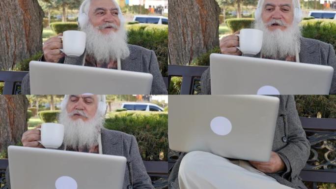 时尚聪明的高级男子拿着杯子喝热饮料，正在使用笔记本电脑和白色无线耳机在视频链接上聊天。现代高级祖父通