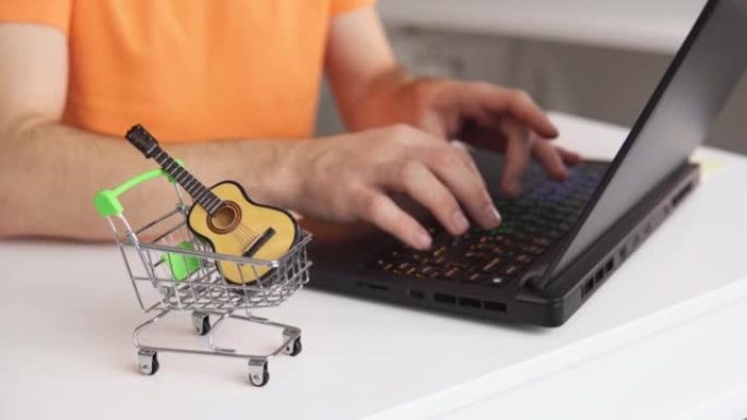 桌子上有微型原声吉他的小型购物车。男子使用笔记本电脑在线购买音乐乐器。