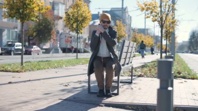 年轻的盲人，智能手机坐在城市公园的长凳上，打电话给