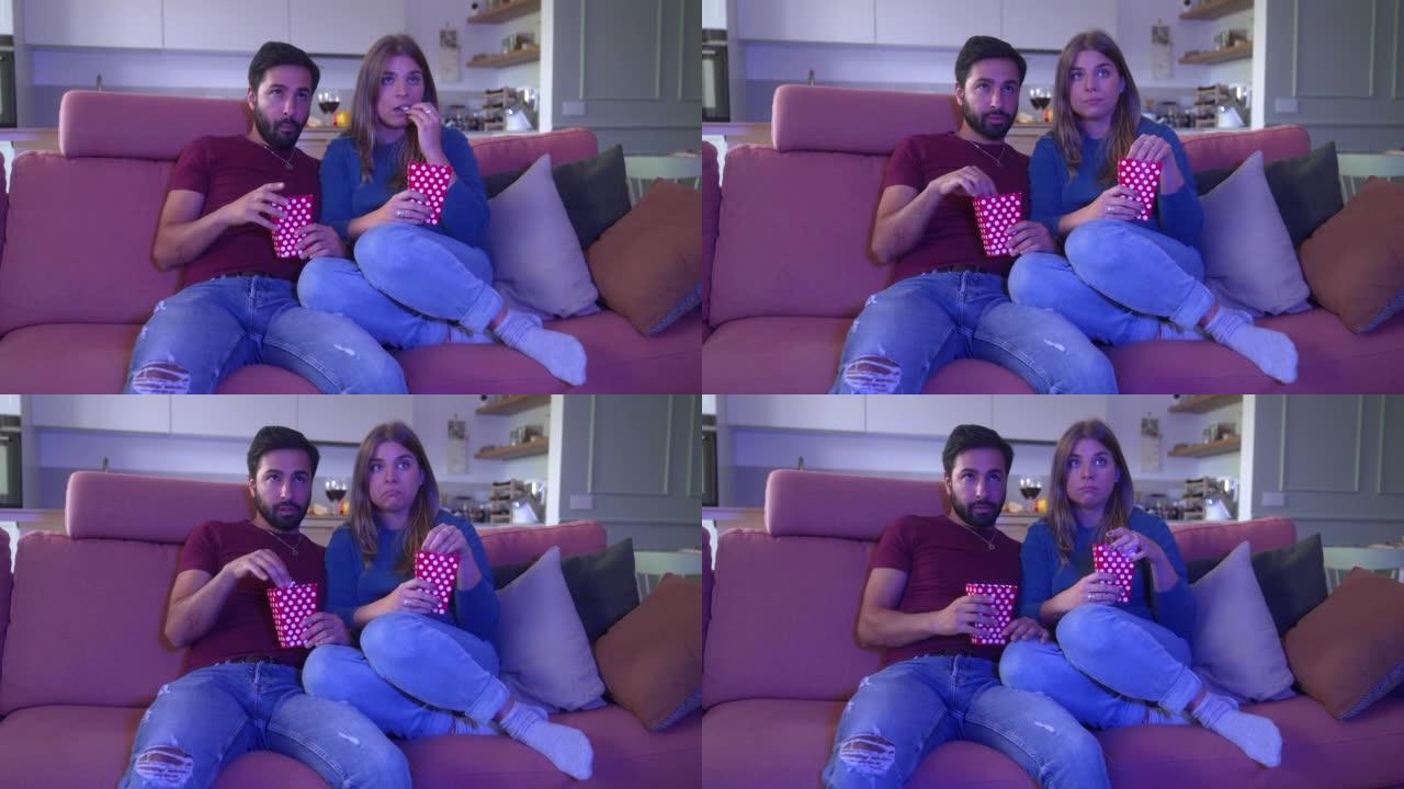 年轻的异性恋夫妇在电视上看恐怖电影，爆米花坐在公寓的沙发上。害怕的夫妇在家里一起在电视上观看可怕的视