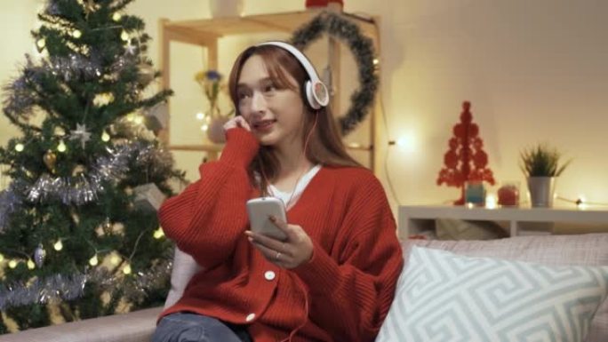 美丽的女士独自坐在为圣诞节装饰的舒适客厅中，拿着智能手机，欣赏耳机中的歌曲。