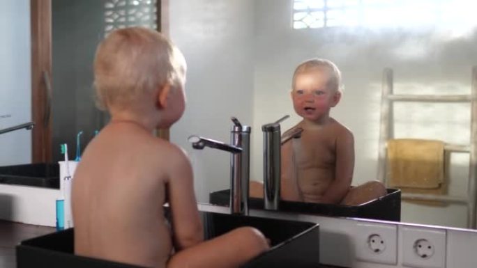 一个可爱的金发碧眼的白人小孩坐在一个方形的黑色设计师水槽里，一边看着镜子里的自己，一边用水玩儿童游戏