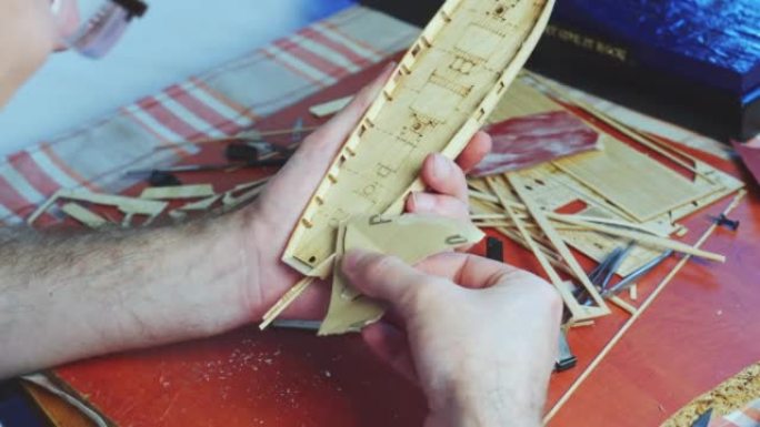 人手调整船模的胶合板细节，在砂纸上打磨。建造玩具船、爱好和手工艺品的过程。工作台有各种材料、零件和工