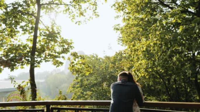 在风景背后，年轻可爱的夫妇在秋天公园的栏杆附近紧密地呆在一起，拥抱，享受愉快的聊天。