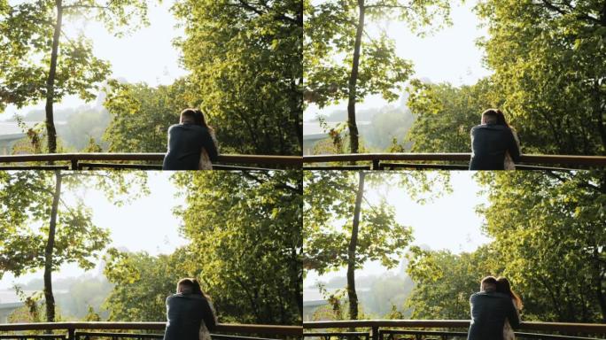 在风景背后，年轻可爱的夫妇在秋天公园的栏杆附近紧密地呆在一起，拥抱，享受愉快的聊天。
