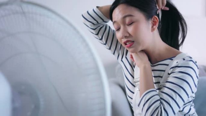 年轻的不快乐的亚洲妇女遭受夏天的高温，并从家里客厅沙发上的风扇中流出空气