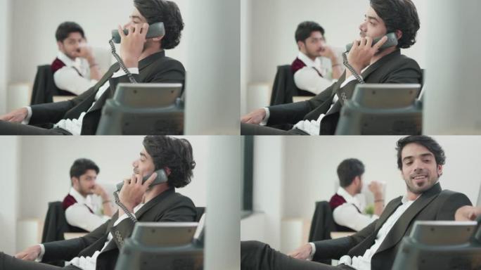 快乐放松的商人微笑着与同事交谈。坐在椅子上，在办公室的桌子上拿着通话固定电话。