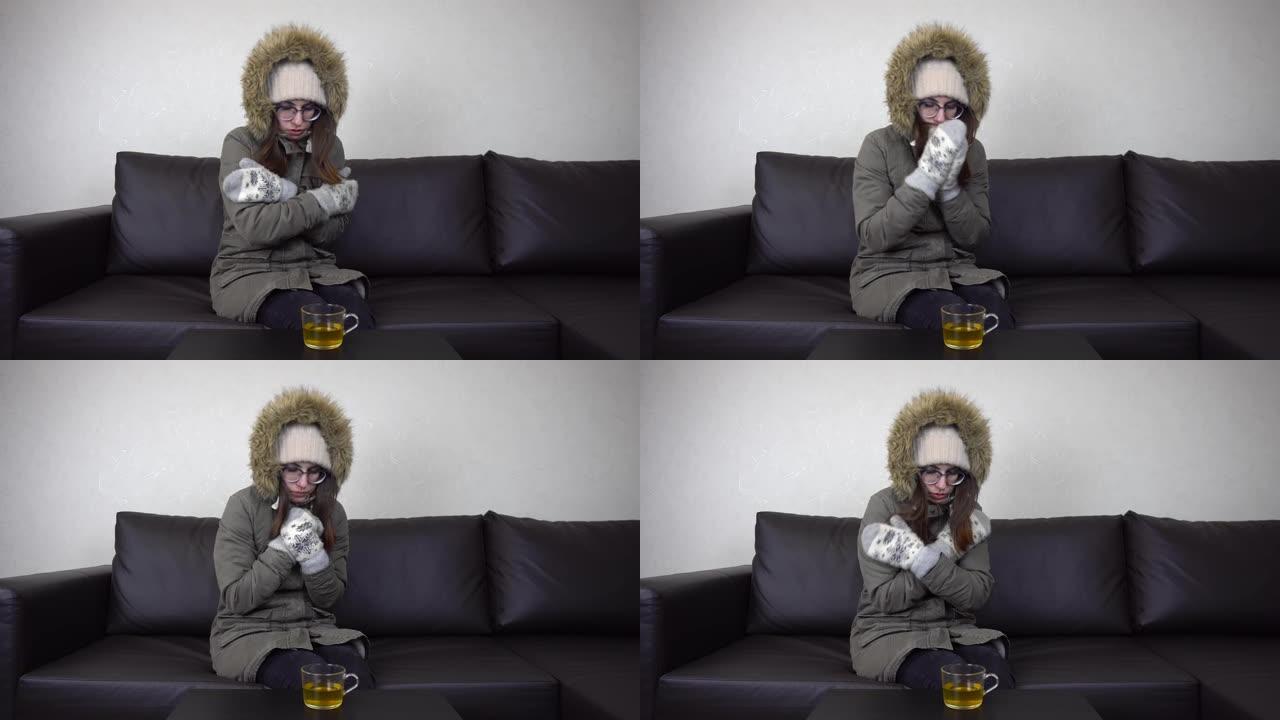 一个年轻的女孩穿着冬衣坐在寒冷的公寓里的沙发上。一个戴眼镜的女人试图通过拥抱自己来取暖。4 k
