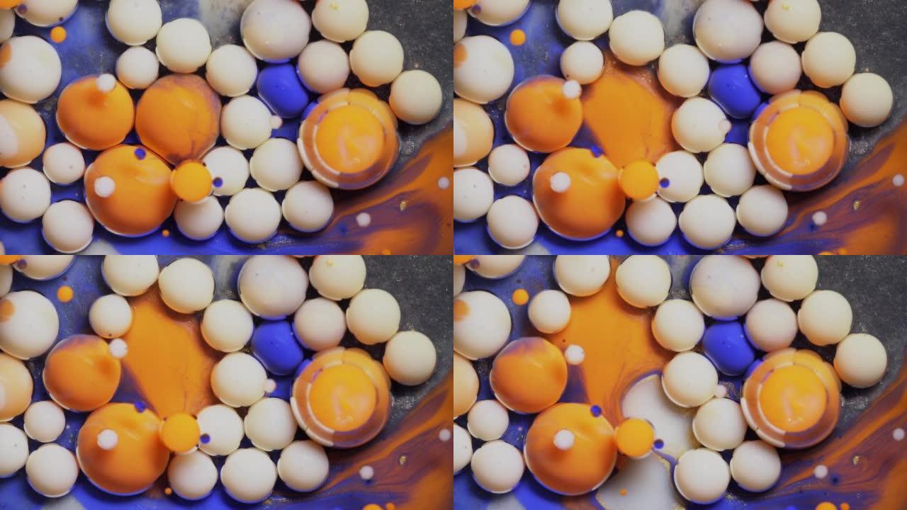特写多色的蓝色橙色和白色气泡在宇宙色彩表面破裂并混合在抽象设计中。金色闪闪发光的颗粒、墨球滴和混合