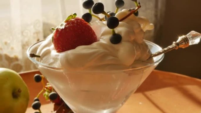 在玻璃冰激凌碗里用新鲜草莓和装饰性蓝色葡萄的奶油冰淇淋特写镜头，用银勺从日落的光线中发光。水果浆果冰