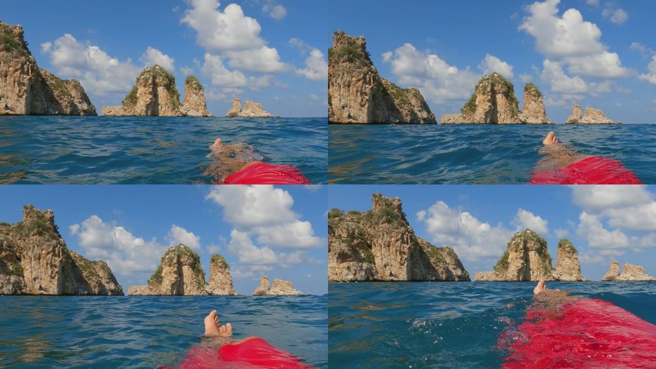 Fpv的男性腿和脚放松，同时漂浮在海水上，背景是Scopello Stacks或Faraglioni
