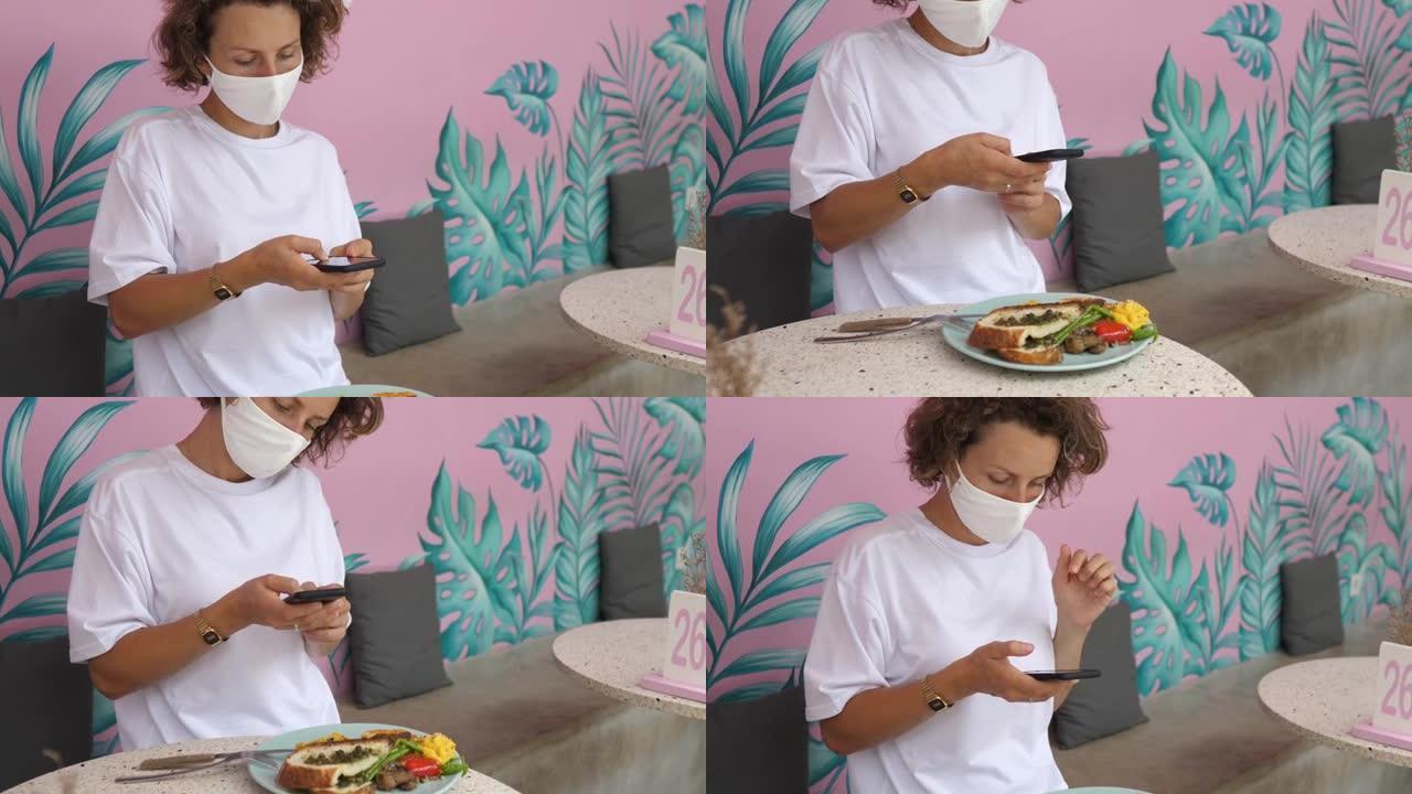 一个脸上戴着防护面具的女孩坐在咖啡馆里，为她订购的午餐食物拍照。她将准备好的菜的照片上传到社交网络。