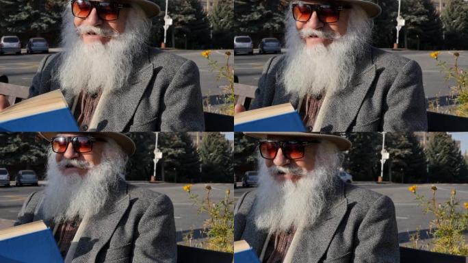 可爱的笑着的祖父戴着帽子，戴着墨镜，留着白色蓬松的胡须，在户外城市的长凳上看书。一个开朗的高年级男子