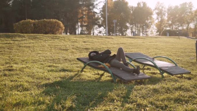 男人躺在秋天阳光明媚的公园的日光躺椅上，戴上兜帽。