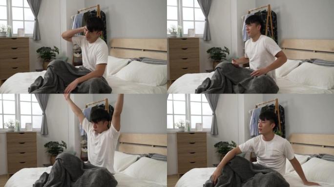 昏昏欲睡的亚洲男性坐在床上，早上在明亮的卧室室内起床后，伸懒腰挠头。
