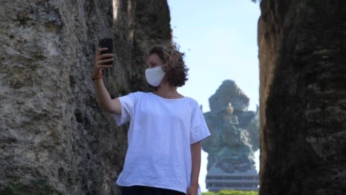 独自去异国他乡旅行。一名戴着病毒面具的年轻女子站在粪便之间，在巴厘岛神庙前自拍，然后回头。