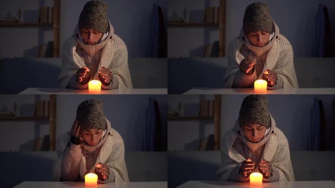一个穿着温暖的冬帽和围巾的男人坐在家里的桌子旁，用燃烧的蜡烛温暖双手。家里冬天不供暖的概念。