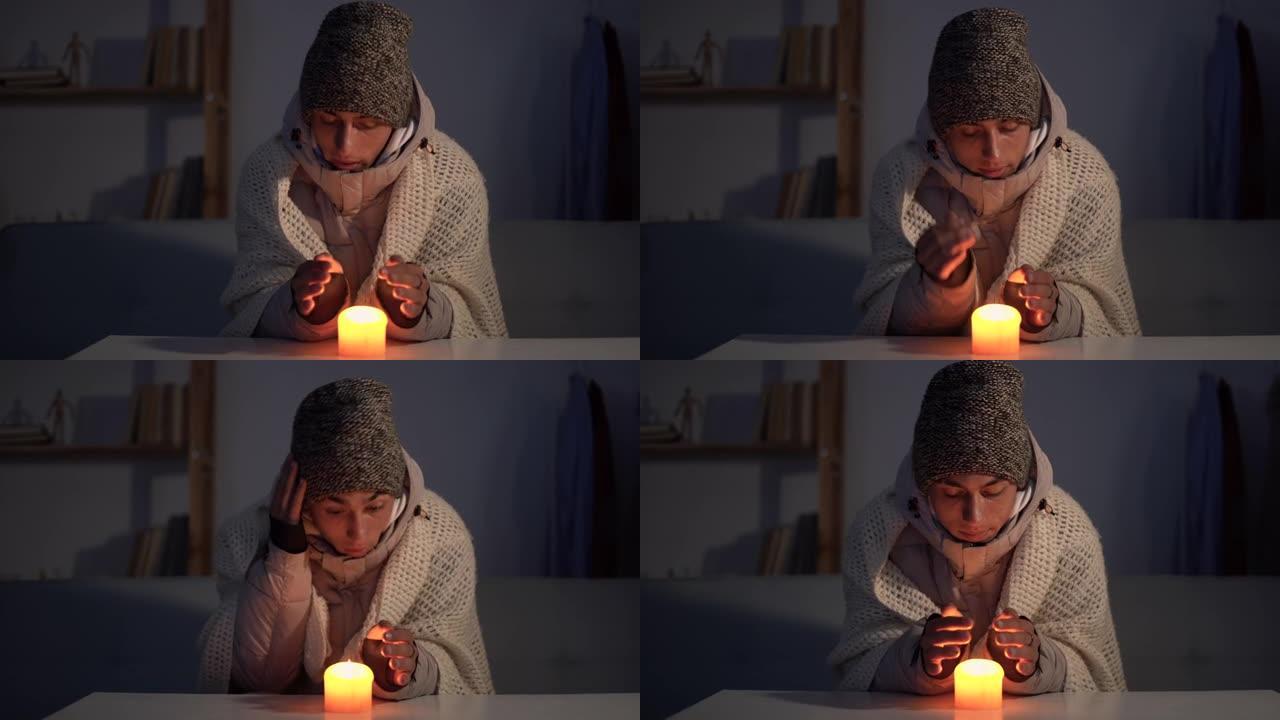 一个穿着温暖的冬帽和围巾的男人坐在家里的桌子旁，用燃烧的蜡烛温暖双手。家里冬天不供暖的概念。