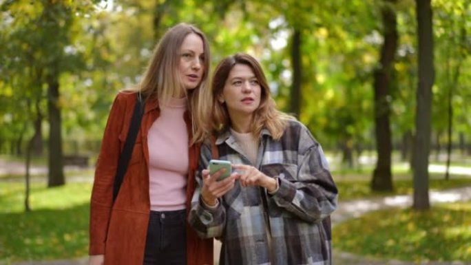 两名自信的白人女性在智能手机上使用在线地图应用程序在秋季公园离开的肖像。美丽苗条的朋友选择方向路线在