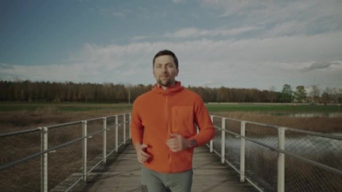 秋天，穿着橙色运动衫的男子慢跑者在斯堪的纳维亚半岛的自然木桥上奔跑。运动员在寒冷的天气里穿过人行天桥