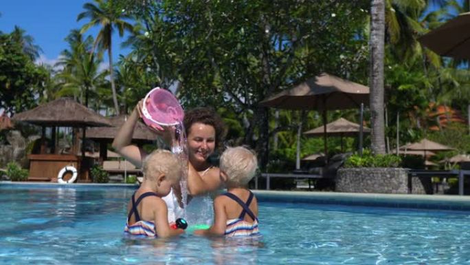 母亲和孩子们一起在游泳池里玩游泳玩具。妈妈把水桶放在头上，从里面倒水，模仿瀑布。假期让孩子们忙碌的小