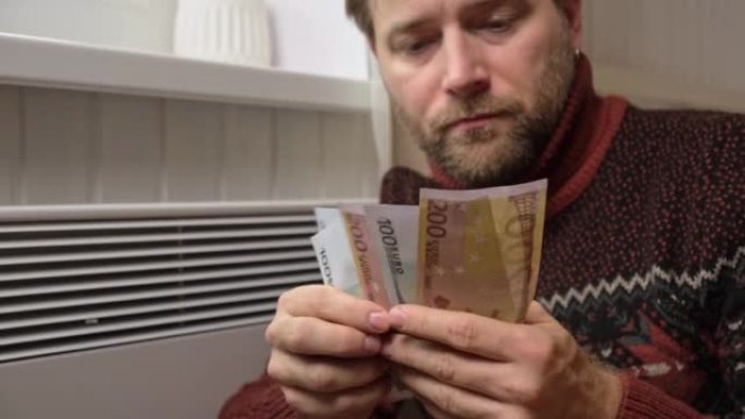 在家中电加热器散热器附近的人手拿着欧元钞票。男人付钱取暖。加热昂贵。节能。生活成本上升。全球天然气危