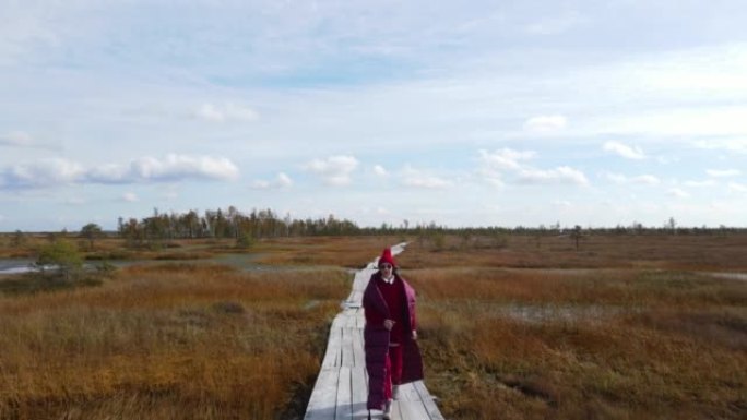在白俄罗斯的叶尔尼亚，一名穿着红毛衣、戴着帽子、戴着围巾的时尚女子在秋天的沼泽中沿着木板铺成的小径走