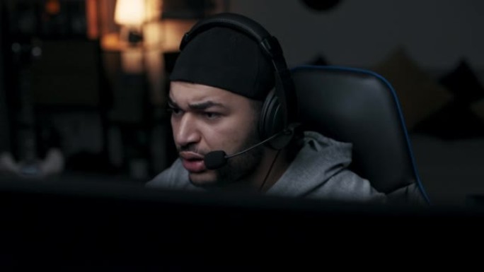 年轻人坐在电脑桌旁的黑暗房间里。穿着灰色t恤的球员头上戴着黑色帽子。男人戴着耳机和游戏伙伴聊天。紧张