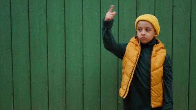 身穿黄色背心，戴着针织帽的男孩孩子在村庄绿色木栅栏的背景下，双手指向左侧。