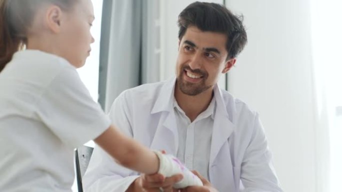 从正面看儿科医生男医生穿着制服咨询小女孩病人，断手裹着白色石膏绷带。