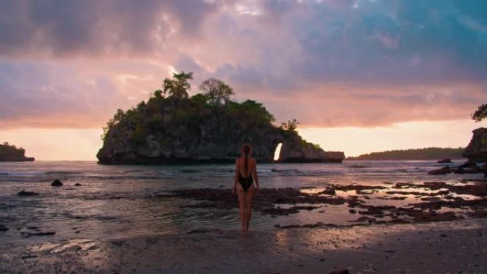 山岛水晶湾海滩上五颜六色的日落天空努沙佩尼达背景后面的泳装孤独瘦女人享受海浪。水面反射的橙色光。