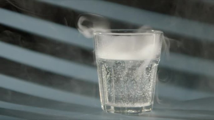 烟来自一杯清水或苏打水。玻璃里有白烟的一杯水。蓝色背景，阳光透过百叶窗。冷却饮料的慢动作视频。