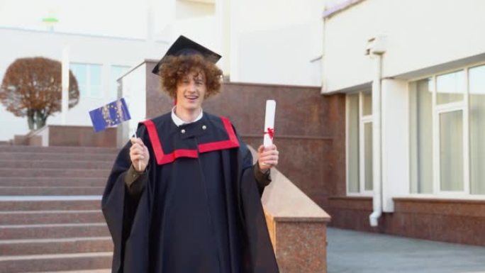一个年轻的卷发有趣的硕士毕业生拿着完成学业的文凭和欧盟旗帜