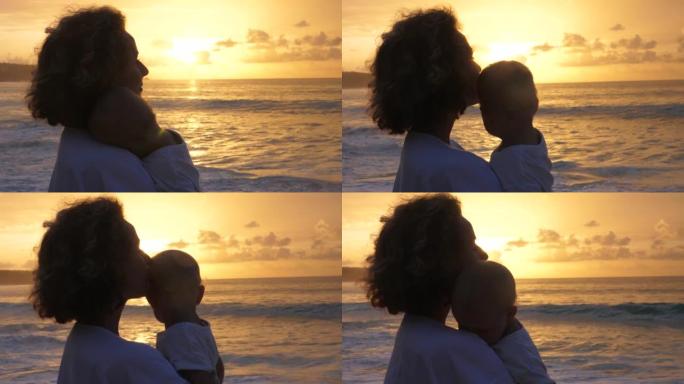母性的快乐。一位年轻的高加索母亲将她的小孩抱在怀里，深情地亲吻他的额头，站在海边，看着日落。
