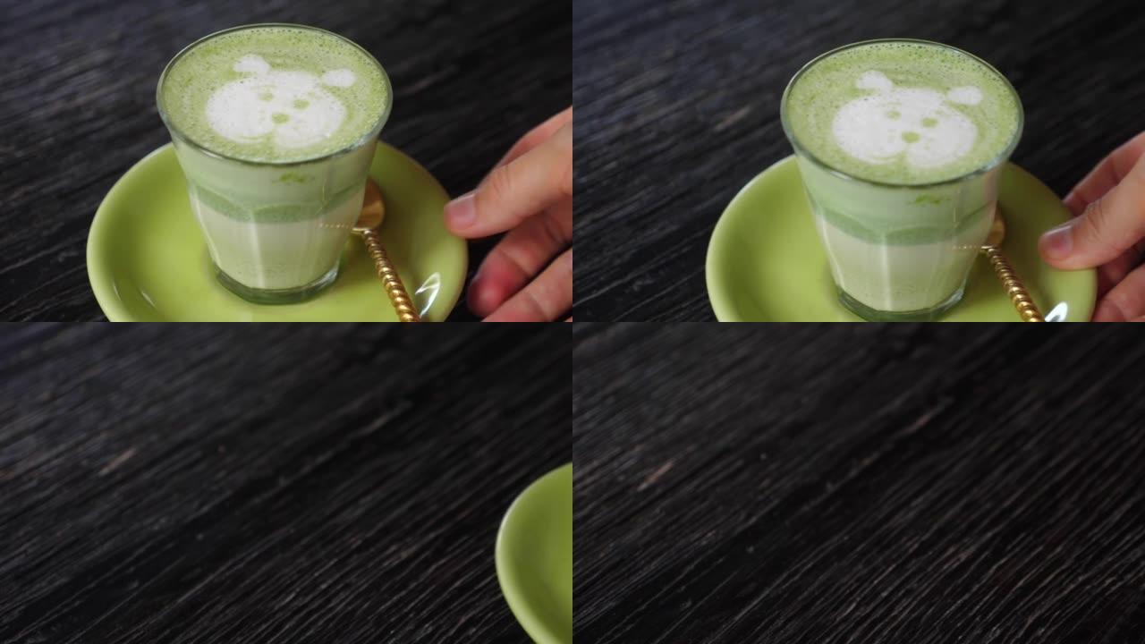一只手将一杯抹茶拿铁茶从桌子上移到绿色的碟子上，并在白色泡沫上发出熊的枪口的欢快图像。美味的食物和饮