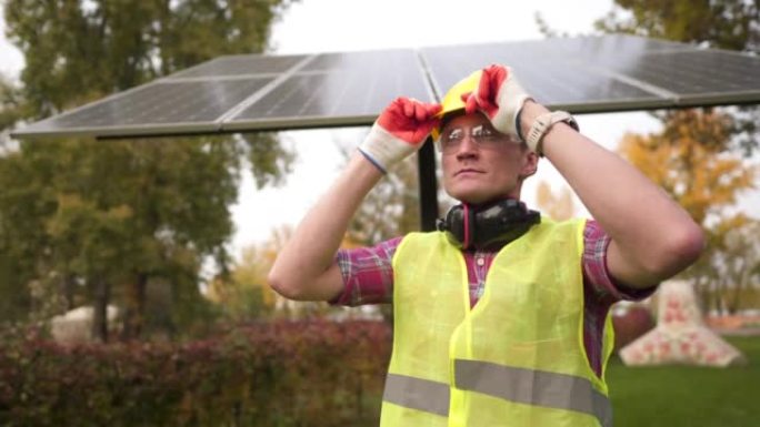 男性太阳能发电厂工程师在太阳能电池板前戴着防护安全帽，穿着防护服。穿着制服的高加索人维护着生产清洁能