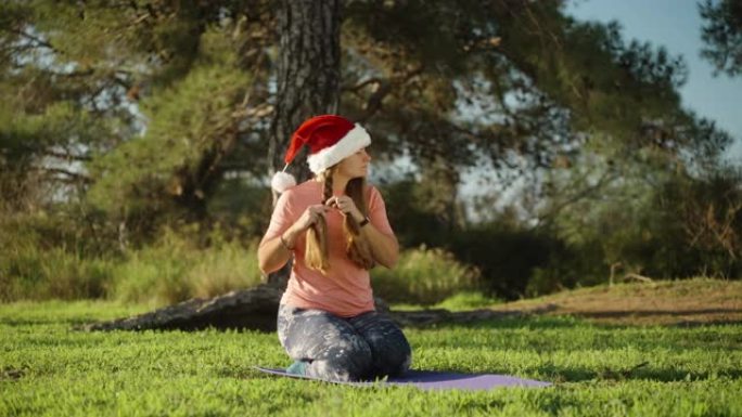 黎明时分，一个戴着红色圣诞老人帽子的森林女孩，坐在瑜伽垫上，将头发编成辫子。