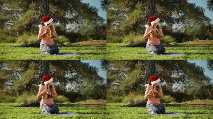 黎明时分，一个戴着红色圣诞老人帽子的森林女孩，坐在瑜伽垫上，将头发编成辫子。