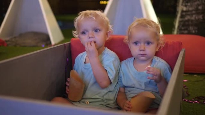 两个可爱的两岁金发碧眼的双胞胎，正坐在室外电影院的软席上，怀着好奇的心情在大屏幕上观看电影，一边吃着