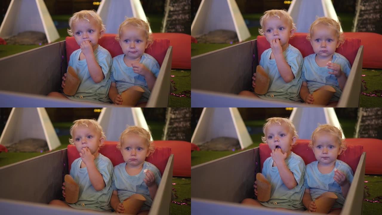 两个可爱的两岁金发碧眼的双胞胎，正坐在室外电影院的软席上，怀着好奇的心情在大屏幕上观看电影，一边吃着