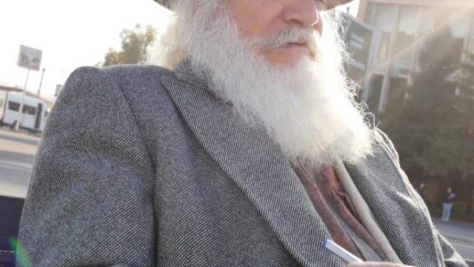 在阳光明媚的日子里，一位戴着帽子和外套的白发大胡子的老年教授坐在户外的长凳上，在笔记本上写了一张纸条