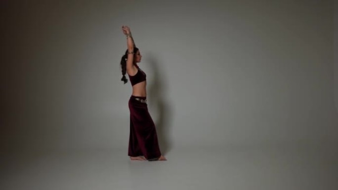侧视图手臂上有翅膀纹身的苗条女人举手慢动作在灰色背景下表演。高加索才华横溢的肚皮舞演员跳舞中东舞蹈。