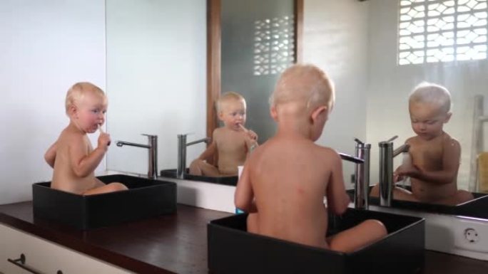 两个高加索双胞胎婴儿各自坐在自己的水槽里，他们像母亲教他们的那样玩水刷牙。他们互相看和复制。