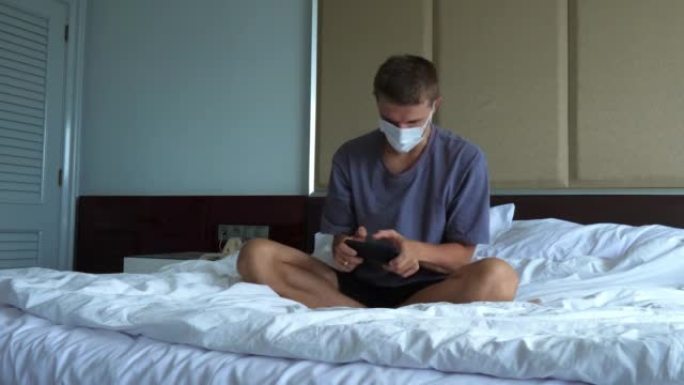 在锁定期间，戴着防护面具的年轻成年男子在床上的智能手机上积极玩电子游戏。健康人和无症状感染患者的隔离