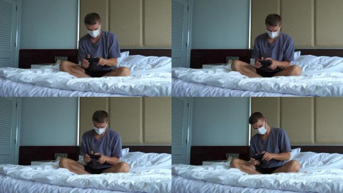 在锁定期间，戴着防护面具的年轻成年男子在床上的智能手机上积极玩电子游戏。健康人和无症状感染患者的隔离